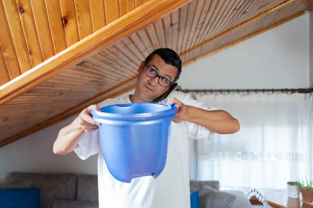 Homeowner catching ceiling drip in a bucket while calling insurance about a roof leak